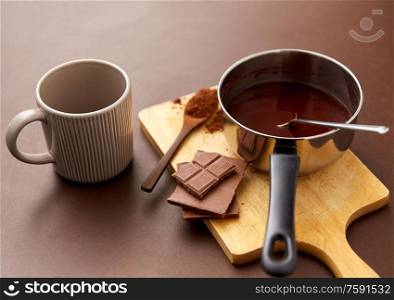 sweets, confectionery and culinary concept - pot with melted hot chocolate, cocoa powder in spoon, ceramic mug and wooden board on brown background. pot with hot chocolate, mug and cocoa powder