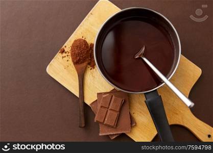 sweets, confectionery and culinary concept - pot with melted hot chocolate, cocoa powder in spoon and wooden board on brown background. pot with melted hot chocolate and cocoa powder