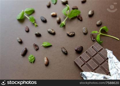 sweets, confectionery and food concept - dark chocolate bar in foil wrapper with peppermint and cocoa beans on brown background. dark chocolate bar with peppermint and cocoa beans