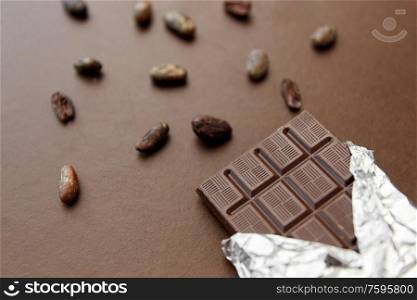 sweets, confectionery and food concept - dark chocolate bar in foil wrapper and cocoa beans on brown background. dark chocolate bar in foil wrapper and cocoa beans