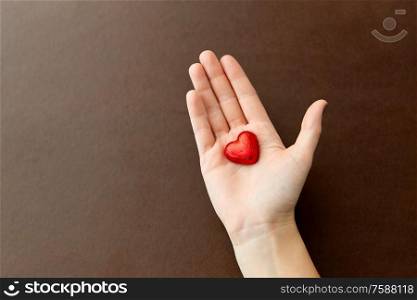 sweets, confectionery and food concept - hand holding heart shaped chocolate candy in red foil wrapper on brown background. hand with red heart shaped chocolate candy