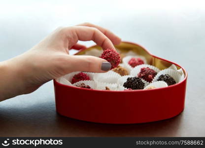 sweets, confectionery and food concept - hand with candies in red heart shaped chocolate box on brown background. hand with candies in heart shaped chocolate box