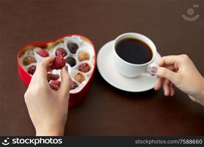 sweets, confectionery and food concept - hands with candies in red heart shaped chocolate box and cup of coffee on brown background. hands, candies in heart shaped box and coffee cup