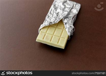 sweets, confectionery and food concept - white chocolate bar in foil wrapper on brown background. white chocolate bar in foil wrapper on brown