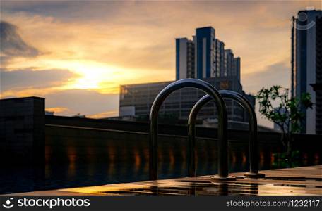 Swimming pool at luxury hotel and spa in the morning with golden sunrise sky. Closeup grab bars ladder at edge of swimming pool. Leisure at hotel poolside concept. Cityscape of skyscraper building.