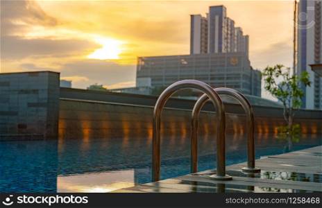 Swimming pool at luxury hotel and spa in the morning with golden sunrise sky. Closeup grab bars ladder with clean water and wooden floor at edge of swimming pool. Leisure at hotel poolside concept.