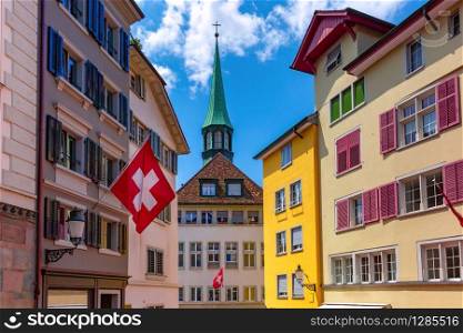 Swiss flag waving, church and colorful facades of houses in Old Town of Zurich, the largest city in Switzerland in sunny day.. Zurich, largest city in Switzerland