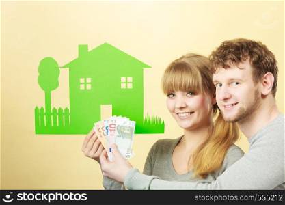 Symbolism property ownership finances mortgage savings concept. Wife and husband holding items. Young couple shows cash with house cutout.. Wife and husband holds paper house.