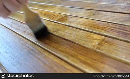 Szene zeigt eine Hand mit einem Pinsel, der eine Flussigkeit auf einen Holztisch auftragt - Fruhjahr oder Herbst, Gartenmobel mussen mit Lasur geschutzt werden.