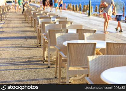 Tables and chairs of the beach cafe are located along the promenade where vacationers walk.. The tables and chairs of the beach cafe are located along the promenade amid strolling people and the blue sea.