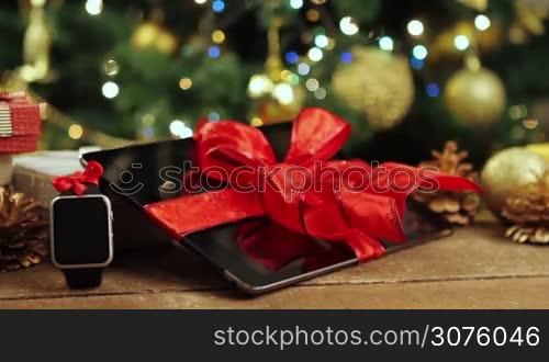 Tablet pc, smartphone and smartwatch with gifts and decorations in front of Christmas tree with lights on wooden table.