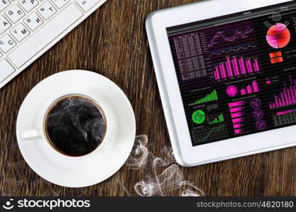 Tablet pc with graphs and cup of coffee on wooden table. Work place