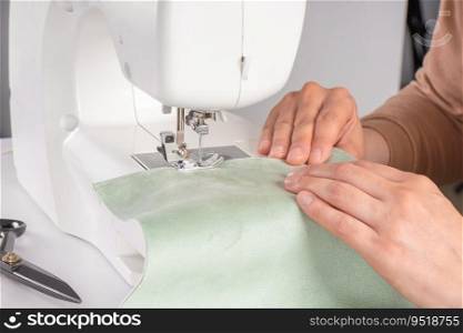 Tailor hands stitching green fabric on modern sewing machine at workplace in atelier. Women’s hands sew pieces of fabric on sewing machine closeup. Handmade, hobby, repair, small business concept