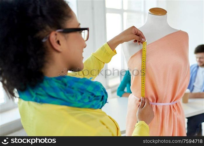 tailoring, sewing and clothing concept - close up of african american fashion designer measuring dress with tape measure. fashion designer measuring dress with tape measure