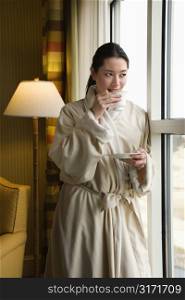 Taiwanese mid adult woman in bathrobe drinking coffee and looking out window.