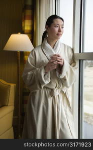 Taiwanese mid adult woman in bathrobe looking out window holding coffee cup.