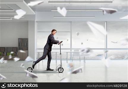 Take a five minute break. Young cheerful businessman in suit riding scooter in office
