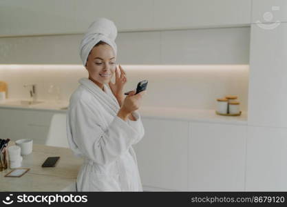 Take care of yourself. Young happy woman in bathrobe looking with smile in compact mirror and applying skin care product on face while standing in modern kitchen and doing cosmetic procedures at home. Happy healthy woman in bathrobe applying skin care product on face after taking shower or bath at home