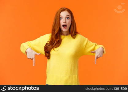 Take look quick. Amazed and fascinated cute ginger girl, redhead student in yellow sweater, gasping open mouth from surprise and wonder, pointing fingers down, look impressed, orange background.. Take look quick. Amazed and fascinated cute ginger girl, redhead student in yellow sweater, gasping open mouth from surprise and wonder, pointing fingers down, look impressed, orange background