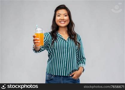 takeaway drinks and people concept - happy smiling young asian woman drinking orange juice from plastic cup with paper straw over grey background. asian woman with juice in plastic cup with straw