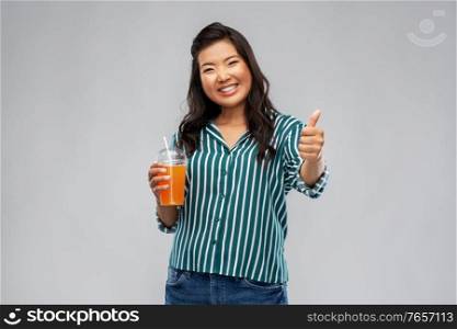 takeaway drinks and people concept - happy smiling young asian woman drinking orange juice from plastic cup with paper straw showing thumbs up over grey background. asian woman with juice in plastic cup with straw