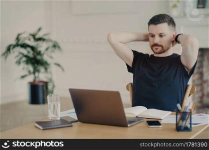 Taking a break. Male freelancer or entrepreneur in relaxed pose wearing casual clothes watching video or read information on laptop computer while working from home or modern office space. Male entrepreneur looking at laptop screen and relaxing while working in modern office