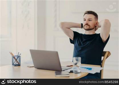 Taking a break. Male freelancer or entrepreneur in relaxed pose wearing casual clothes watching video or read information on laptop computer while working from home or modern office space. Young businessman enjoying work done while sitting at his workplace with laptop