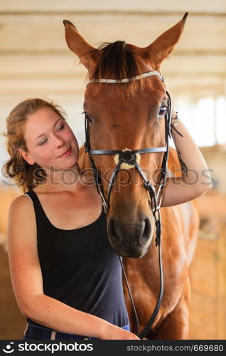 Taking care of animals, love and friendship concept. Jockey young girl petting and hugging brown horse in stable. Jockey young girl petting and hugging brown horse