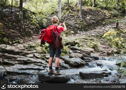 Taking pictures from vacation. Woman with backpack taking photos of landscape using smartphone camera standing on rock on mountain stream during summer trip