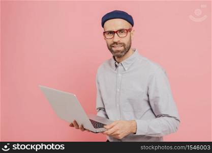 Talented male journalist with appealing look, holds modern laptop computer, works on new article, dressed in fashionable clothes, models against pink background, connected to wireless internet
