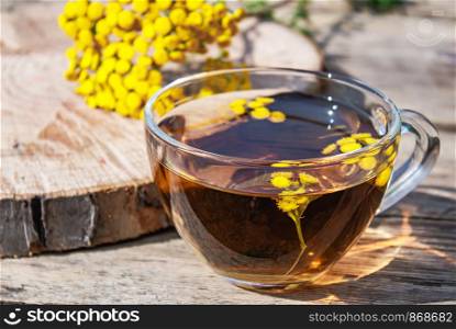 Tansy infusion in a glass cup and yellow tansy flowers on a wooden table. Tansy Herbal tea. Healing herbs. Phytotherapy.. Tansy infusion in a glass cup and yellow tansy flowers on a wooden table. Tansy Herbal tea. Healing herbs.