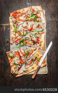 tarte flambee mit paparika,arugula and creme fresh with kitchen knife on rustic wooden background, top view