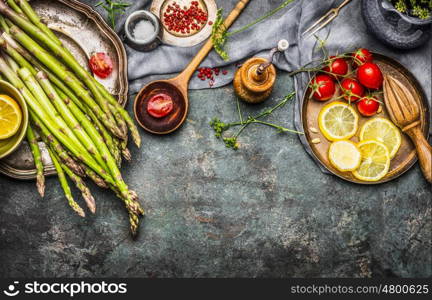 Tasty asparagus cooking with tomatoes, lemon and seasoning, preparation on rustic kitchen table with wooden spoon, top view, border. Dark style