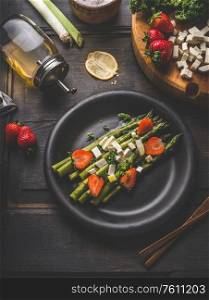 Tasty asparagus salad topped with strawberries and fresh cheese cubes served in black plate on dark rustic table background with ingredients. Top view. Healthy food. Home cooking and eating