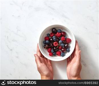 Tasty berries, freshly picked fruits on ceramic plate in the female hands on a concrete background. Concept of healthy food with copy space. Flat lay. Natural organic fruits, berries in a plate on a gray stone background. Women&rsquo;s hands hold white bowl with cherry, raspberry, blueberries.