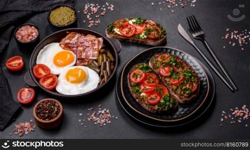 Tasty breakfast consists of eggs, bacon, beans, tomatoes, with spices and herbs on a dark concrete background. Tasty breakfast consists of eggs, bacon, beans, tomatoes, with spices and herbs