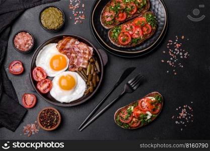Tasty breakfast consists of eggs, bacon, beans, tomatoes, with spices and herbs on a dark concrete background. Tasty breakfast consists of eggs, bacon, beans, tomatoes, with spices and herbs