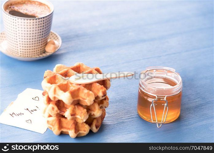 Tasty breakfast with fresh waffles with honey, a hot cup of coffee and a cute paper note with the message missing you, under the morning light, on a blue table.