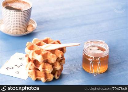 Tasty breakfast with fresh waffles with honey, a hot cup of coffee and a cute paper note with the message missing you, under the morning light, on a blue table.