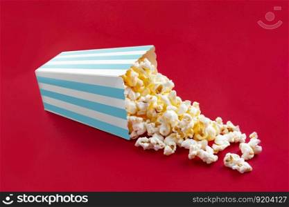 Tasty cheese popcorn falling out of a turquoise striped carton bucket, isolated on red background. Scattering of popcorn grains. Movies, cinema and entertainment concept.