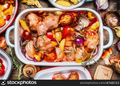 Tasty chicken with colorful vegetables and red sweet paprika in casserole, preparation on rustic wooden background with bowls and cutting cooking ingredients, top view, close up