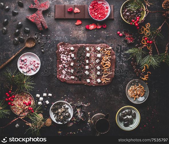 Tasty Christmas chocolate bark with topping of caramel, marshmallows, nuts and candy . Christmas sweet gifts preparation with ingredients on dark background with copy space. Top view