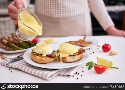 Tasty eggs Benedict - woman pours hollandaise sauce onto poached eggs.. Tasty eggs Benedict - woman pours hollandaise sauce onto poached eggs