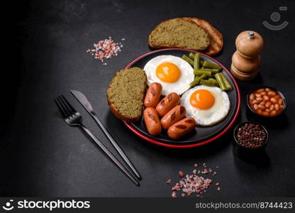 Tasty English breakfast of fried eggs, beans, asparagus, sausages with spices and herbs on a dark concrete background. Tasty English breakfast of fried eggs, beans, asparagus, sausages with spices and herbs