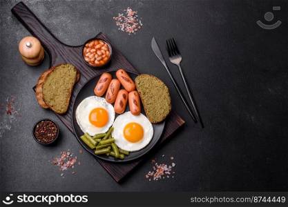 Tasty English breakfast of fried eggs, beans, asparagus, sausages with spices and herbs on a dark concrete background. Tasty English breakfast of fried eggs, beans, asparagus, sausages with spices and herbs