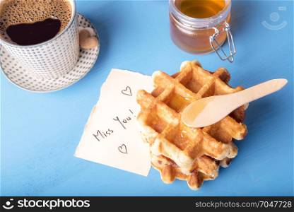 Tasty fresh waffles with honey on top, a cup of hot coffee and a cute note on a piece of paper with the miss you message and hearts, on a blue wooden table.