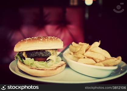 Tasty hamburger and french fries on plate in american food restaurant. Red leather sofa in the background. Vintage, retro style