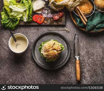 Tasty homemade burger on plate with knife and ingredients on dark rustic background, top view