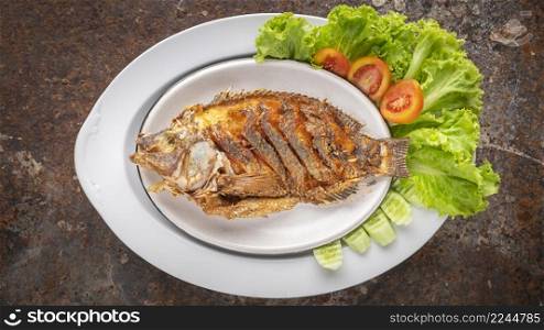 tasty large fried nile tilapia fish with lettuce, tomato and cucumber in ceramic plate on rusty texture background, top view