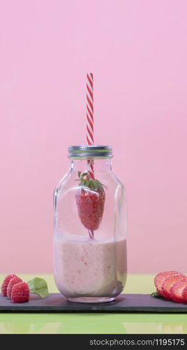 Tasty looking milkshake decorated with a straw piercing a strawberry inside a glass jar bottle on a dark plate with raspberries and strawberry slices on a pink background. Healthy and vegetarian food concept.. Tasty looking strawberry milkshake in a glass bottle jar